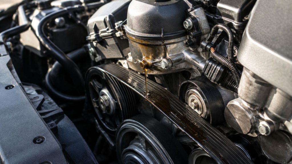 BMW engine with an oil filter housing leak dripping oil onto the serpentine belt, illustrating how oil contamination can damage belt performance and lead to engine repair concerns.